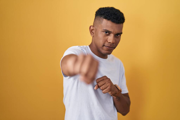 Молодий іспаномовний чоловік стоїть над жовтим тлом, ударяючи кулаком в бій, агресивний і сердитий напад, загроза і насильство
  - Фото, зображення