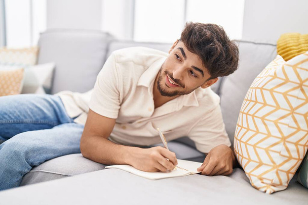 Young arab man writing on notebook sitting on sofa at home - Foto, afbeelding
