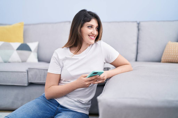 Young hispanic woman using smartphone sitting on floor at home - Foto, Imagen