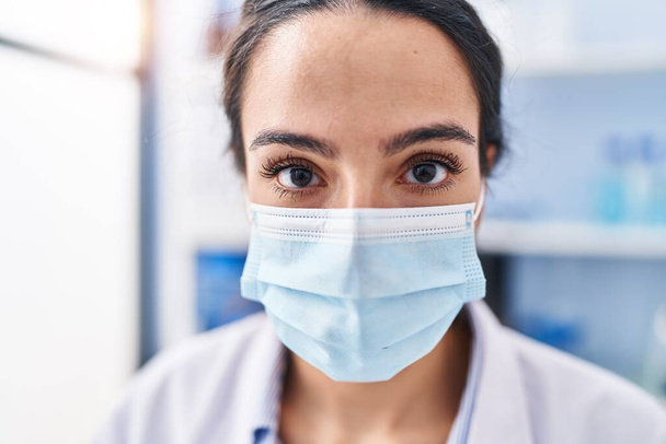Laboratuvarda bilim adamı üniforması ve tıbbi maske giyen genç İspanyol kadın. - Fotoğraf, Görsel
