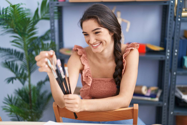 Young hispanic woman artist smiling confident holding paintbrushes at art studio - 写真・画像
