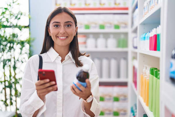 Young beautiful hispanic woman customer using smartphone holding medicine bottle at pharmacy - Fotó, kép