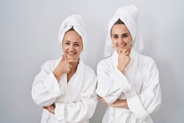 Μέση ηλικία γυναίκα και η κόρη φορώντας λευκό μπουρνούζι και πετσέτα αναζητούν αυτοπεποίθηση στην κάμερα χαμογελώντας με σταυρωμένα χέρια και το χέρι έθεσε στο πηγούνι. θετική σκέψη.  - Φωτογραφία, εικόνα