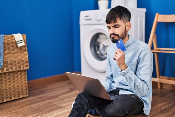 Young hispanic man using laptop and credit card waiting for washing machine at laundry room - Foto, Imagem