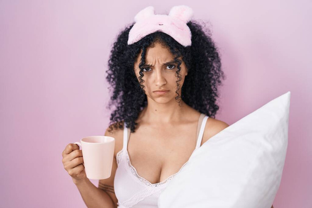 Латиноамериканка з кучерявим волоссям у піжамі п'є чашку кави скептично і нервово, насуплена через проблеми. негативна людина.  - Фото, зображення