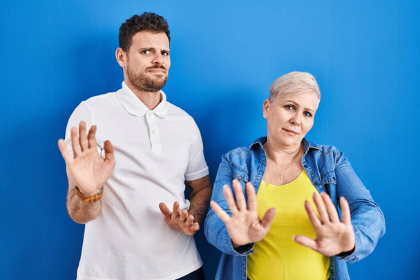 Młoda brazylijska matka i syn stojący nad niebieskim tłem odsuwają dłonie pokazując odmowę i zaprzeczenie ze strasznym i obrzydliwym wyrazem twarzy. zatrzymać i zabronione.  - Zdjęcie, obraz