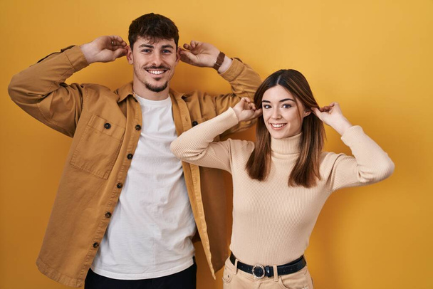 若いヒスパニック系のカップルの黄色の背景に指で耳を引く笑みを浮かべて立って、面白いジェスチャー。オーディション問題  - 写真・画像