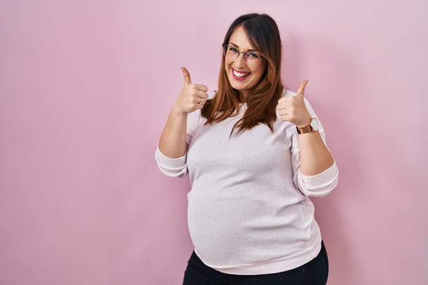 Έγκυος γυναίκα στέκεται πάνω από ροζ φόντο επιτυχία σημάδι κάνει θετική χειρονομία με το χέρι, τους αντίχειρες επάνω χαμογελώντας και χαρούμενος. Χαρούμενη έκφραση και χειρονομία νικητή.  - Φωτογραφία, εικόνα