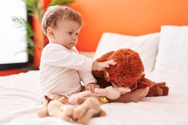 Imádnivaló szőke kisbaba kezében baba ül az ágyon a hálószobában - Fotó, kép