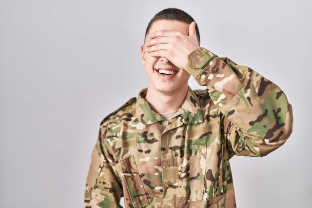 Młody człowiek w mundurze wojskowym uśmiecha się i śmieje z ręką na twarzy, zakrywając oczy niespodzianką. ślepa koncepcja.  - Zdjęcie, obraz