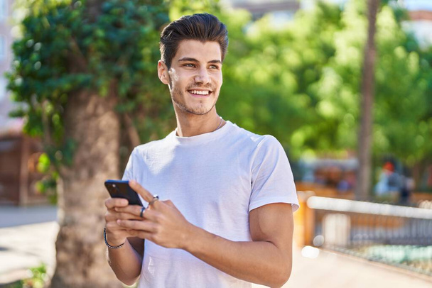 Nuori latino mies hymyilee luottavainen käyttämällä älypuhelin puistossa - Valokuva, kuva