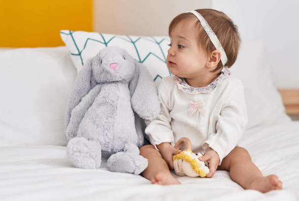 Liebenswertes kaukasisches Baby sitzt auf Bett und spielt mit Puppe im Schlafzimmer - Foto, Bild
