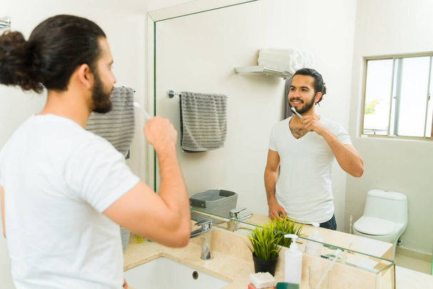 gelukkig jongeman glimlachen kijkend in de badkamer spiegel en poetsen zijn tanden tijdens zijn ochtend routine - Foto, afbeelding