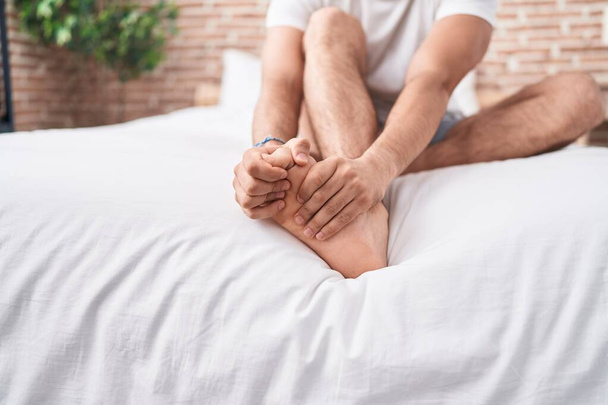 Young hispanic man suffering for feet pain sitting on bed at bedroom - Foto, Imagem