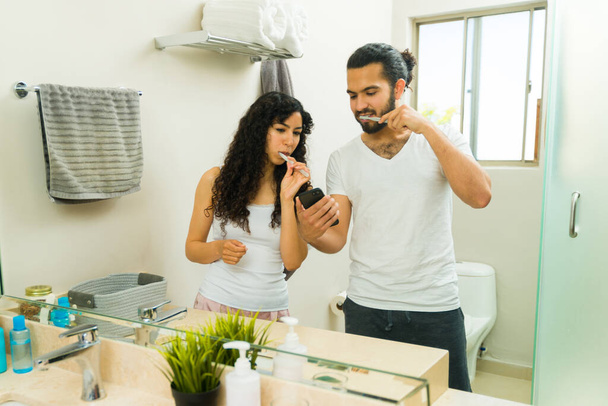 Μεξικάνικο ζευγάρι στο μπάνιο κοιτάζοντας το smartphone ενώ βουρτσίζουν τα δόντια τους μαζί το πρωί - Φωτογραφία, εικόνα