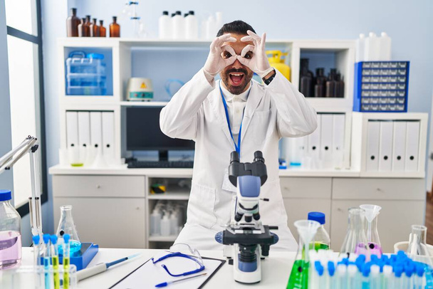 Un joven hispano con barba trabajando en el laboratorio científico haciendo un buen gesto como binoculares sacando la lengua, ojos mirando a través de los dedos. expresión loca.  - Foto, Imagen