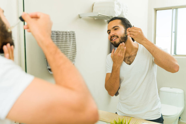 Alegre hombre atractivo usando aceite hidratante en su barba y aseo en el espejo del baño  - Foto, imagen