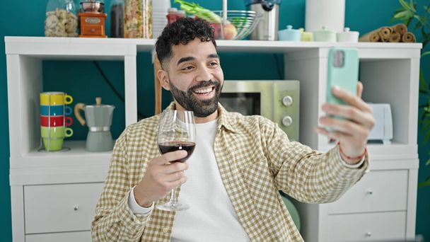 Νεαρός Ισπανός πίνει ένα ποτήρι κρασί και κάνει βιντεοκλήση στην τραπεζαρία. - Φωτογραφία, εικόνα