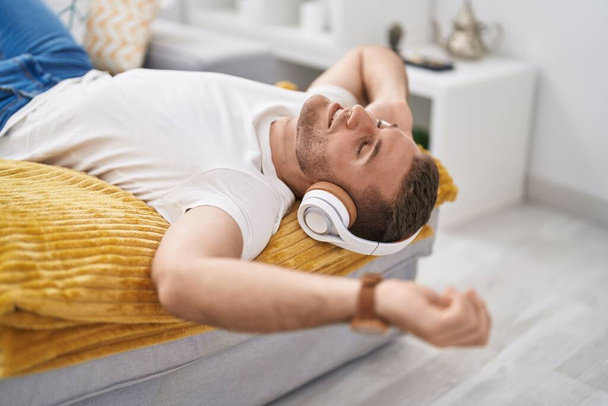 Young caucasian man listening to music lying on sofa at home - Фото, изображение