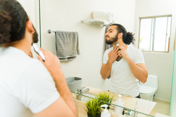 Jongeman trimt zijn baard met behulp van een trimmer tijdens het verzorgen in de ochtend  - Foto, afbeelding