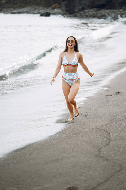 Brunette girl running on the black sand beach wearing a cute bathingsuit in Bali Indonesia. Girl having fun by the ocean - 写真・画像