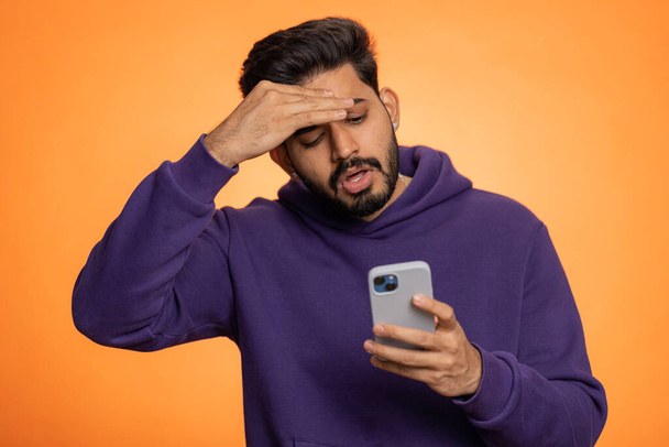 Indian man use mobile cell phone typing browsing, loses becoming surprised sudden lottery results, bad fortune, loss, fail. Handsome hindu guy isolated alone on orange studio wall background, indoors - 写真・画像