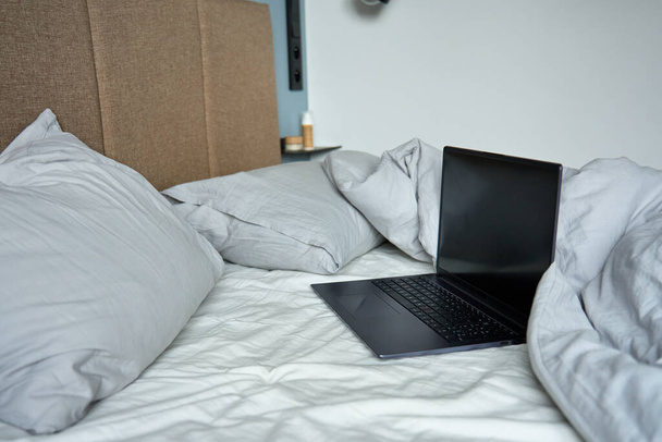 Открыт ноутбук в невыделанной кровати с подушками и измятыми простынями по утрам. Удобное рабочее место для фрилансера, работа на дому - Фото, изображение