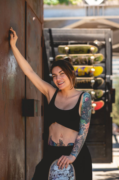 Seksowny miejski styl: portret młodej buntowniczki w czarnej bluzce i szortach, tatuaże i deskorolka w miedzianej ścianie w miejskim skateparku - Zdjęcie, obraz