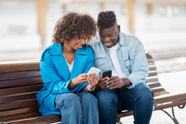 Fröhliches schwarzes Paar, das Smartphones benutzt, während es draußen auf einer Bank sitzt, glückliche junge afroamerikanische Männer und Frauen Wifi teilen oder Fotos auf ihren Mobiltelefonen ansehen, Nahaufnahme - Foto, Bild
