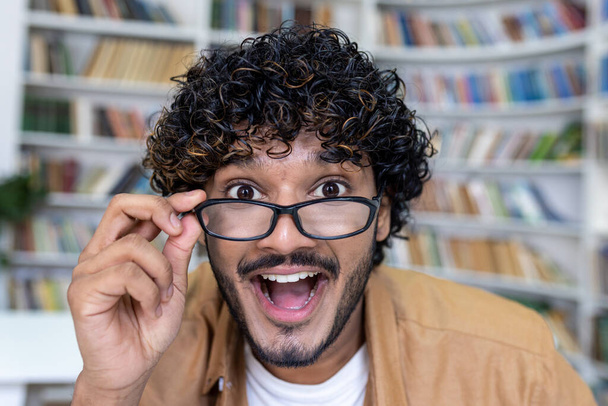 Zaskoczony człowiek patrzący w kamerę, latynoski zbliżenie uśmiechający się i zdejmujący okulary, student wewnątrz biblioteki uniwersyteckiej. - Zdjęcie, obraz