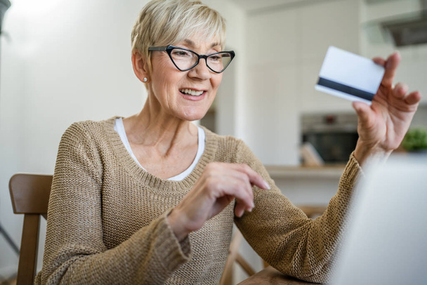 Egy érett idős nő nagymama ül otthon használja hitel vagy betéti kártya online vásárlás böngészés internetes üzletek használja laptop számítógép vásárol dolgokat valós emberek másolni helyet - Fotó, kép