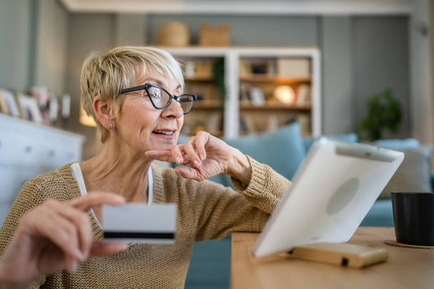 Jedna dojrzała starsza kobieta babcia siedzieć w domu używać karty kredytowej lub debetowej do zakupów online przeglądać sklepy internetowe kupowanie rzeczy korzystać z tabletu cyfrowego prawdziwi ludzie kopiować przestrzeń - Zdjęcie, obraz