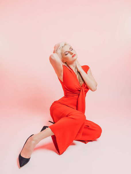 Portrait of a beautiful blonde woman in a red pantsuit.. Posing while sitting in the studio on a pink background. - Φωτογραφία, εικόνα