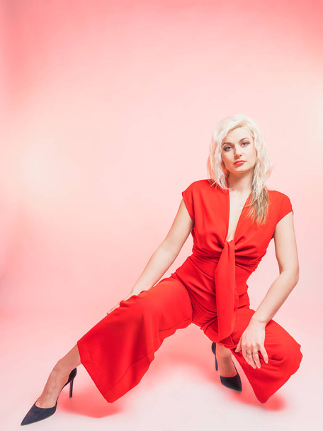 Portrait of a beautiful blonde woman in a red pantsuit.. Posing while sitting in the studio on a pink background. - Foto, Imagem