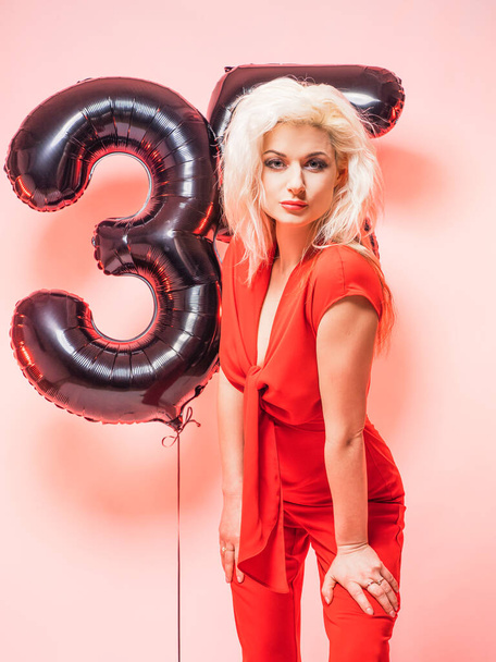 Gyönyörű szőke, piros estélyi ruhában és lufikban 35. Megünnepli a születésnapját. Rózsaszín háttér. - Fotó, kép