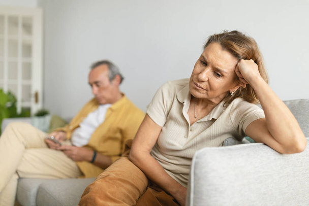 Homem sênior usando smartphone ao ter uma discussão com sua esposa que se sente sozinho, sentado no sofá em casa. Cônjuges não falando depois de uma briga - Foto, Imagem