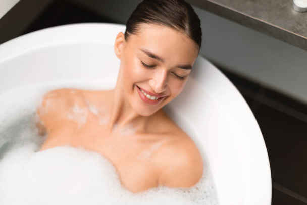 Χαλαρός Lady μπάνιο που βρίσκεται στο νερό με φυσαλίδες αφρού χαλαρώνοντας με τα μάτια κλειστά στο σύγχρονο μπάνιο στο σπίτι. Πάνω άποψη πλάνο της ευτυχισμένης γυναίκας λαμβάνοντας εσωτερικό μπάνιο. Ευεξία και Spa - Φωτογραφία, εικόνα
