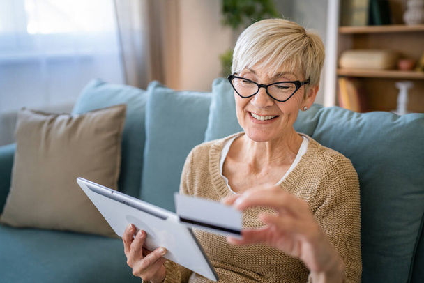 Μια ώριμη ηλικιωμένη γιαγιά κάθεται στο σπίτι χρήση πιστωτικής ή χρεωστικής κάρτας για online αγορές περιηγηθείτε στο διαδίκτυο καταστήματα αγοράζουν πράγματα χρησιμοποιούν ψηφιακό tablet πραγματικό άνθρωποι αντιγράψετε χώρο - Φωτογραφία, εικόνα