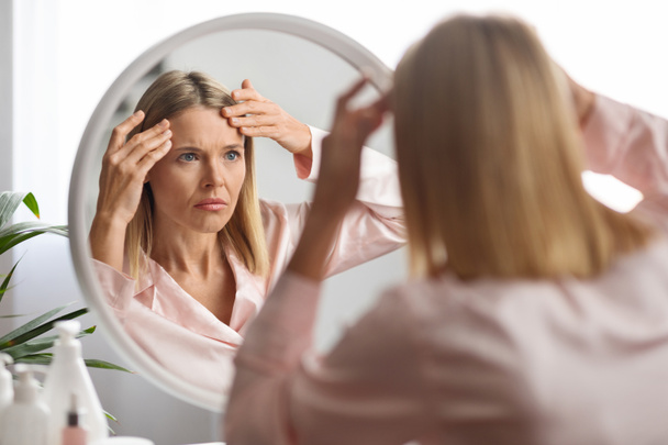 Middle Aged Woman Looking In Mirror And Touching Wrinkles On Her Face, Upset Beautiful Mature Female Examining Fine Lines On Forehead, Suffering Skin Aging, Selective Focus On Reflection, Closeup - Foto, imagen