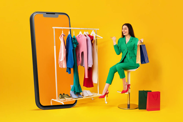 Mobile shopping offer. Shopaholic woman pointing at clothes on clothing rail in huge cellphone, sitting over yellow studio background. E-commerce, fashion and technology concept - Photo, Image