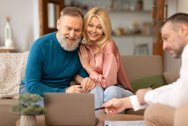Male Consulting Senior Couple Showing Laptop Offering And Selling Real Estate Sitting On Couch At Home. Spouses Talking To Consultant. Internet Service Offer. Selective Focus - Photo, Image