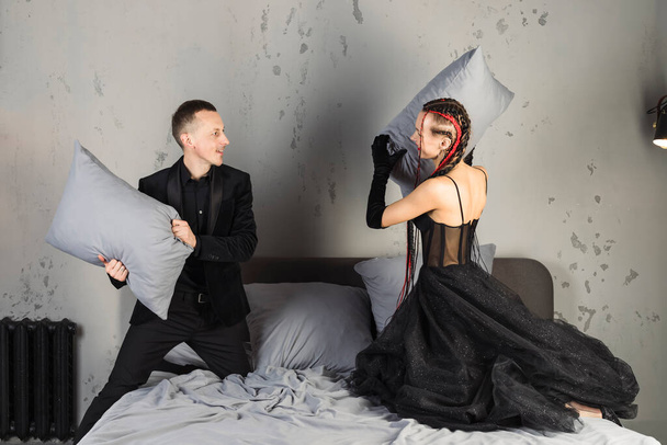 A graceful and elegant woman in stylish black dress with long red braids and man in suit are fighting with pillows. - Photo, image