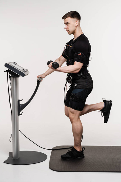 Atleta maschio muscolare con manubri in tuta EMS che utilizza impulsi elettrici per stimolare i muscoli su sfondo bianco. Allenamento sportivo in tuta di stimolazione muscolare elettrica - Foto, immagini