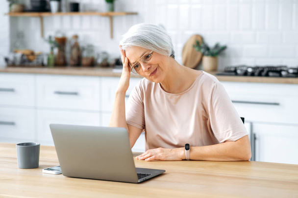 Mujer de mediana edad caucásica insatisfecha con el pelo gris, con gafas, se sienta en una mesa con un ordenador portátil en la cocina en casa, trabaja de forma remota, mira la pantalla en la confusión, lee noticias - Foto, imagen