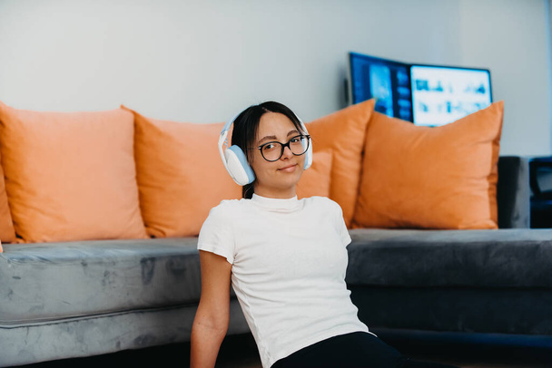 Young woman wearing glasses and headphones while training at home. Her active pose and determination showcase her commitment to a healthy lifestyle, with the modern interior serving as a backdrop for - Photo, Image