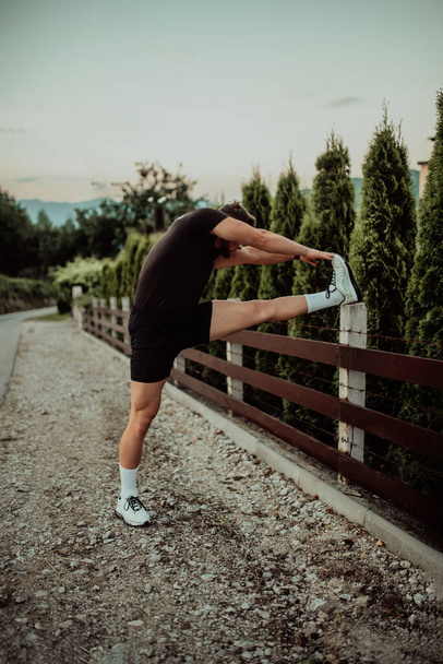 Спортсмен выполняет упражнения на растяжение ног после пробежки в естественной среде. Фотография на тему фитнеса, демонстрирующая гибкость и здоровье физической активности - Фото, изображение