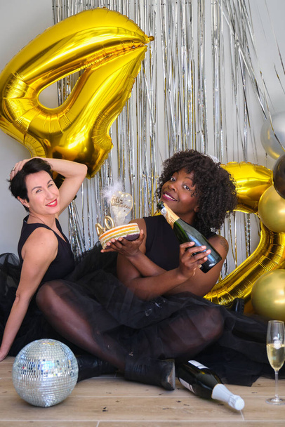 Two multiracial women in a 40 birthday party smiling with champagne bottle and cake. - Fotó, kép