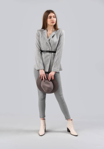 Stylish young woman in jacket and jeans holding hat on grey background - Foto, Imagen