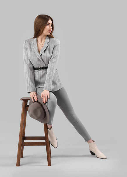 Stylish young woman in jacket and jeans sitting on stool against grey background - Photo, image