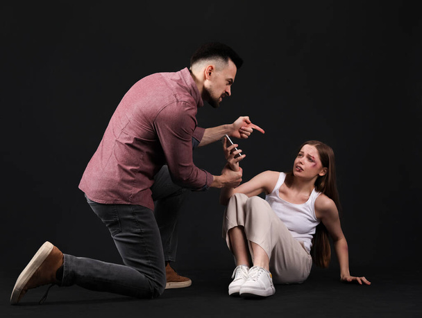 Bruised young woman with mobile phone and her angry husband on dark background. Domestic violence concept - Φωτογραφία, εικόνα